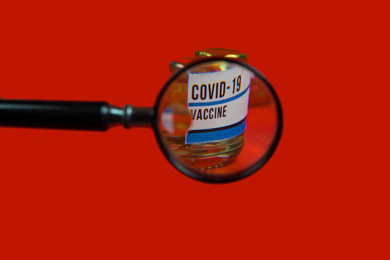 Una lupa aplicada sobre un boto de vacuna covid-19, sobre fondo rojo.