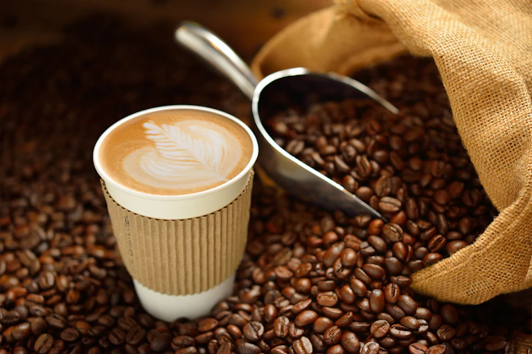 A latte in a disposable cup amid a bag of roasted coffee beans.