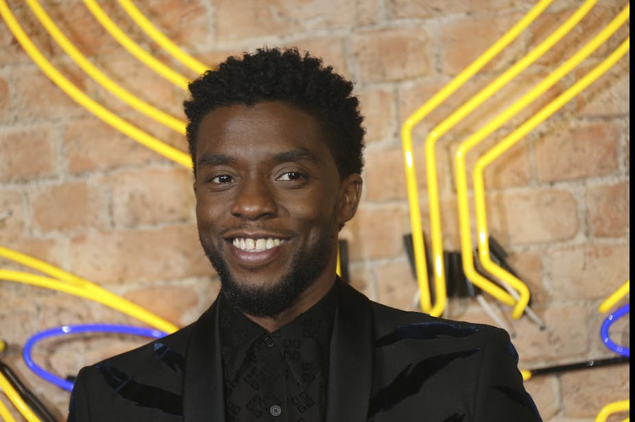 Actor Chadwick Boseman seen from the shoulders up smiling in front of a brick wall with neon lights on it.