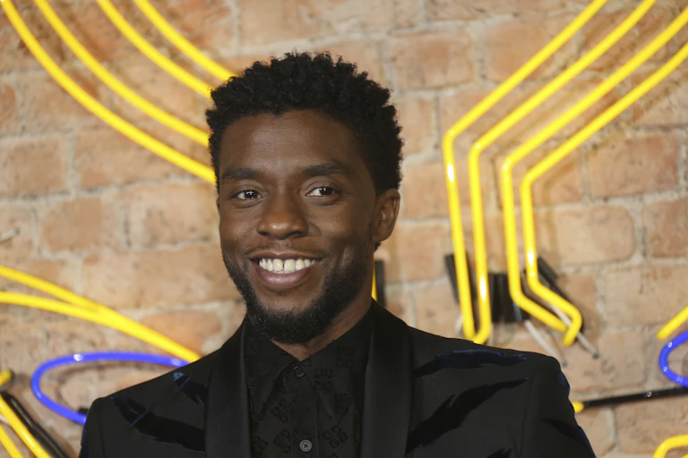 Actor Chadwick Boseman seen from the shoulders up smiling in front of a brick wall with neon lights on it.