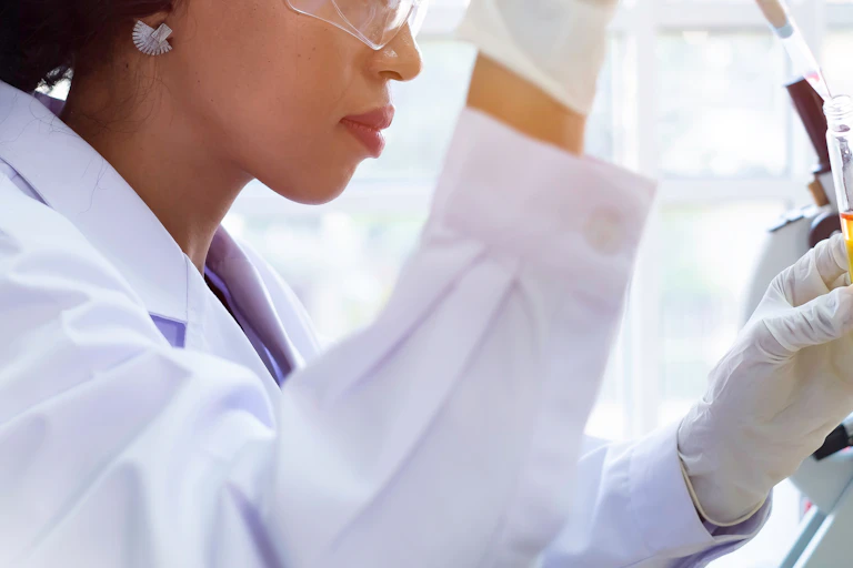 A woman in a lab coat holds a test tube full of liquid alongside a microscope