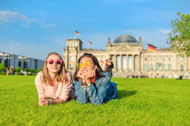Two young women lying on grass in sunshine outside Bundestag building in Berlin.