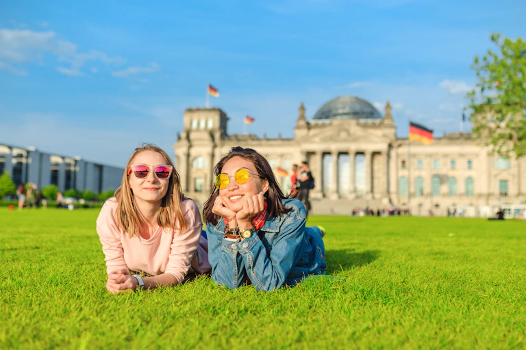 Two young women lying on grass in sunshine outside Bundestag building in Berlin.