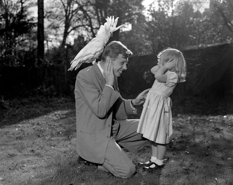 A man with bird on his back kneels beside his daughter.