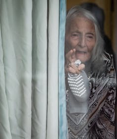 An elderly woman waves from behind a curtain