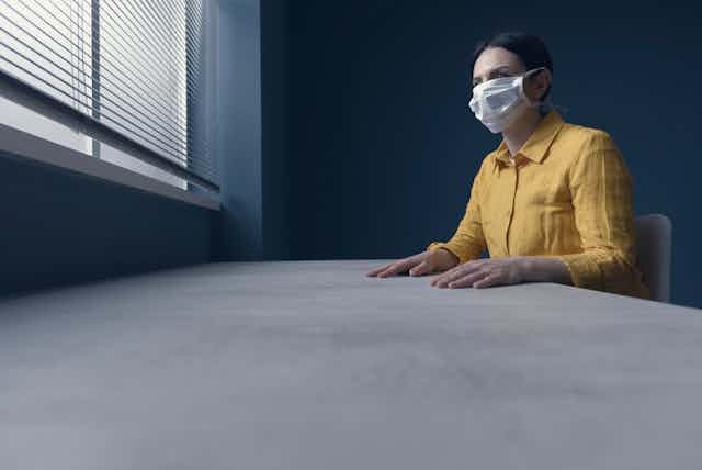 Una mujer con mascarilla en una habitación gris mira hacia la ventana luminosa tapada por una persiana.