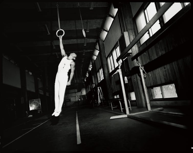 Yukio Mishima, dressed in white, hangs, bloodied, from a still ring.