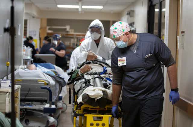 Medical staff push a gurney down a hall where another patient waits in a hospital bed.