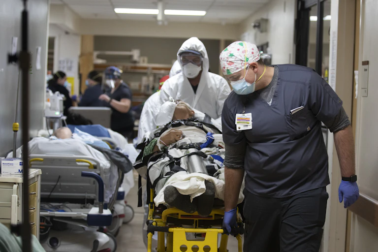 Medical staff push a gurney down a hall where another patient waits in a hospital bed.