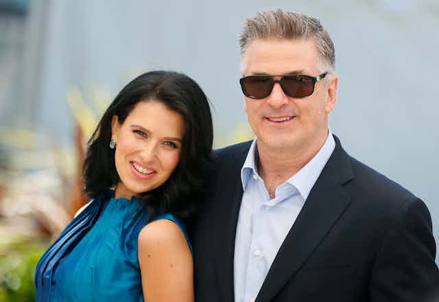 Woman with long dark hair in blue dress alongside man in lounge suit and open-necked shirt wearing sunglasses.