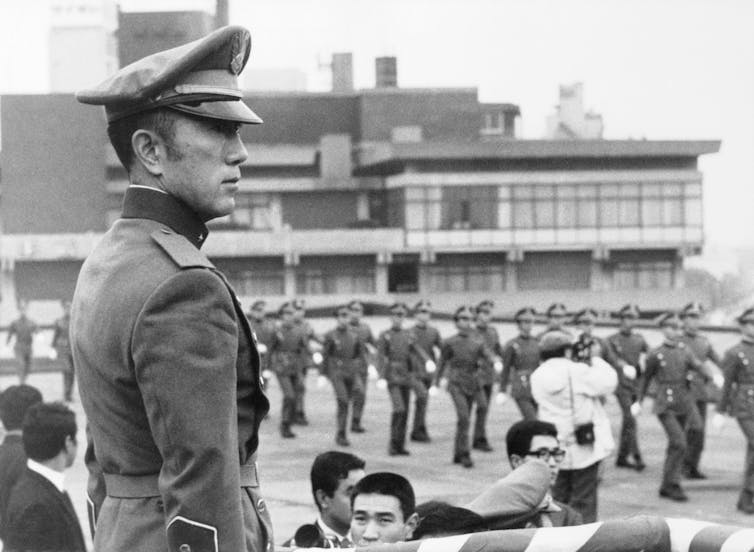 Wearing a military uniform, Mishima watches marchers.