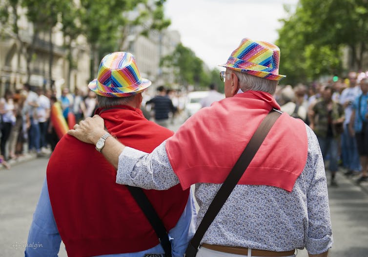 An elderly couple at a Pride parade seen from behind are wearing hats with the colours of the rainbow flag.