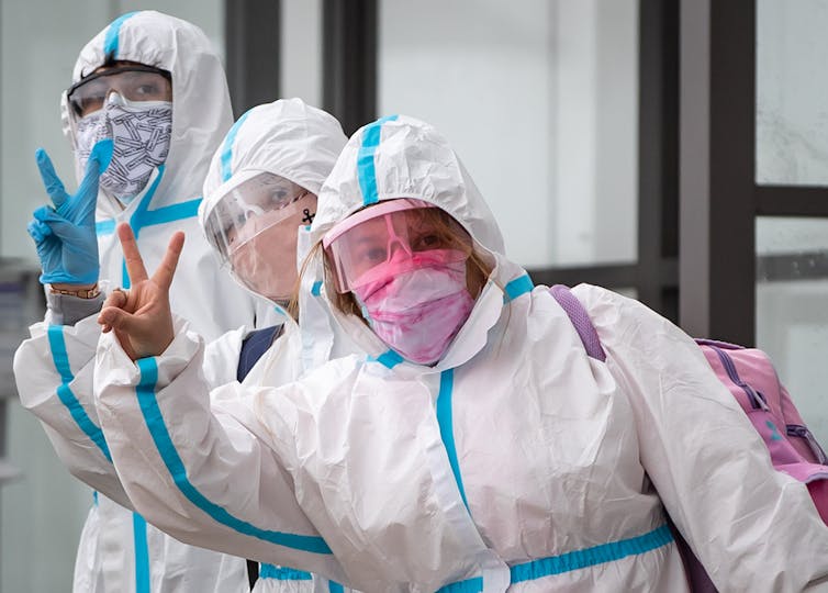 Three travellers in masks, face shields and white haz-mat suits give the peace sign to a photographer.