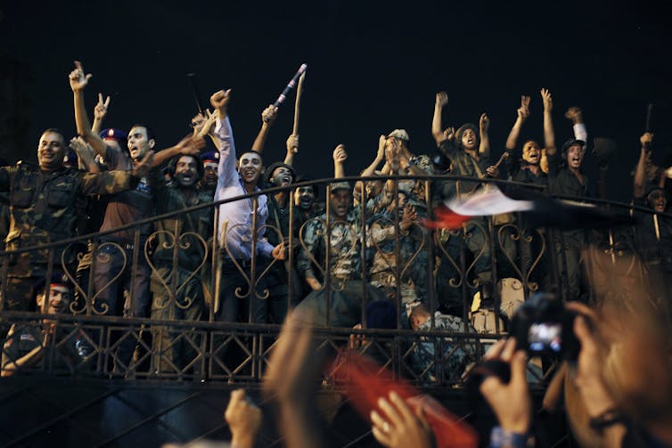 Civilians and soldiers in fatigues holding weapons cheer on a balcony, at night