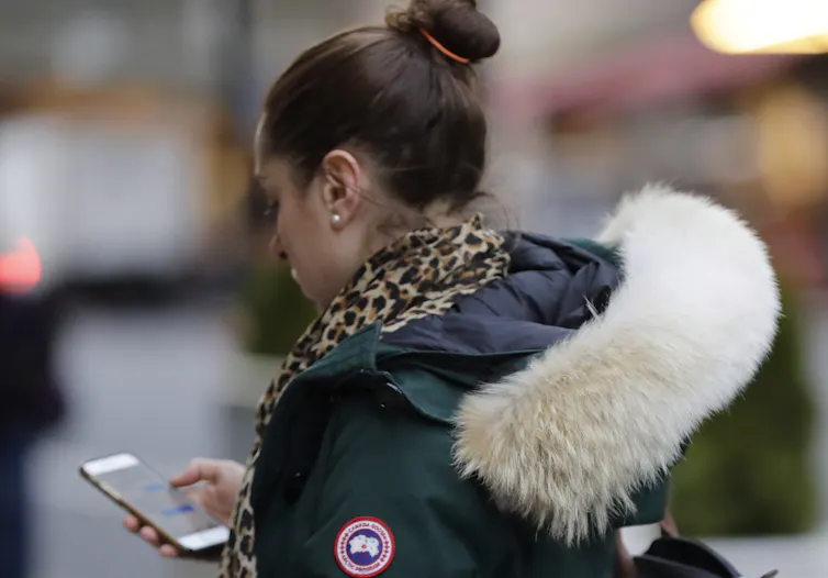 A woman looks at her phone as she walks along a street wearing a Canada Goose coat.