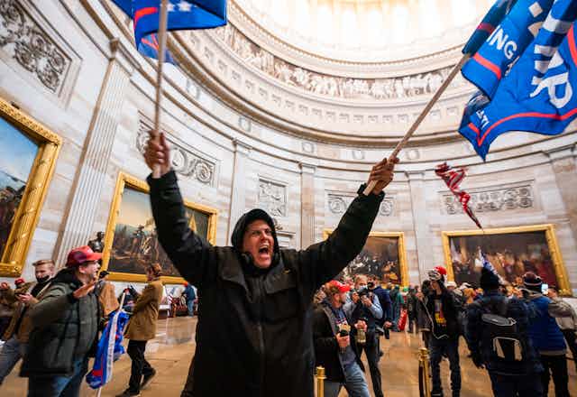 A pro-Trump rioter waving flags and shouting inside the US Capitol building, surrounded by other protesters.