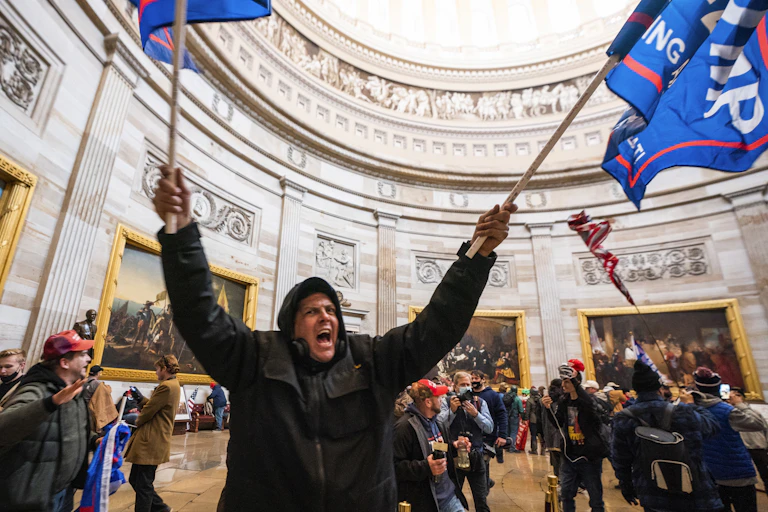 A pro-Trump rioter waving flags and shouting inside the US Capitol building, surrounded by other protesters.