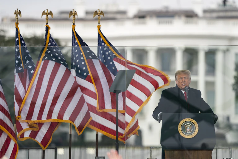 Trump speaks behind plexiglass with the White House in the background.