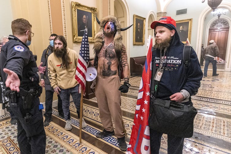 Several protesters, including a shirtless man wearing a fur hat with horns, confront a security guard at the U.S. Capitol.