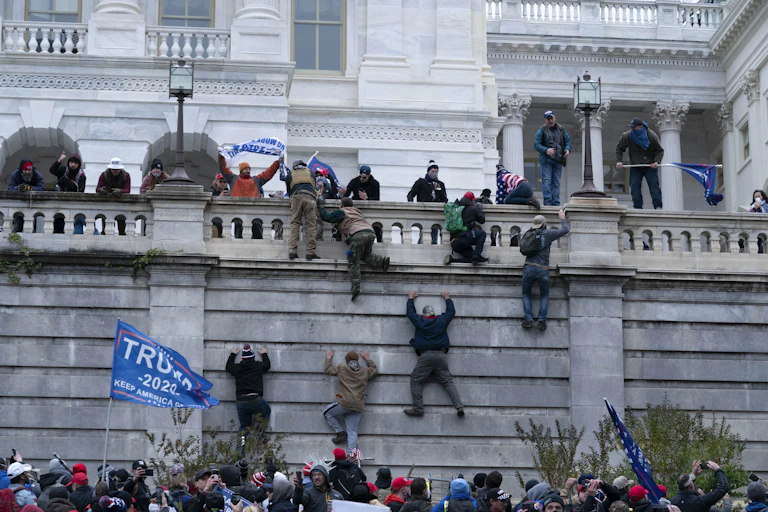 People climb up the wall of the Capitol. Several pro-Trump flags are visible.