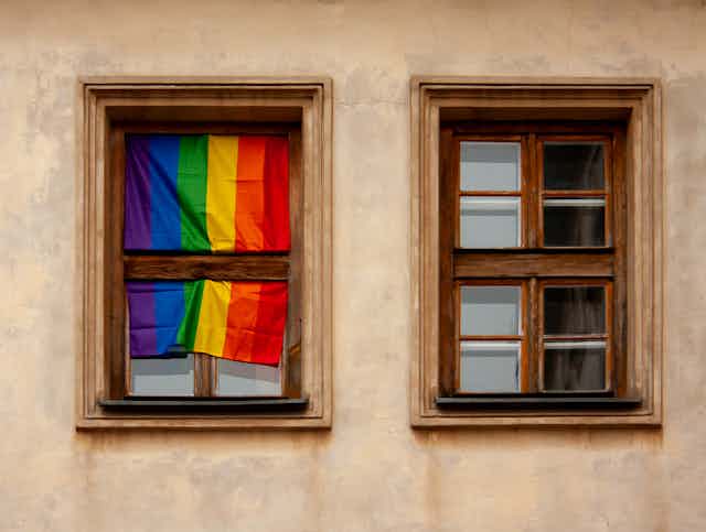 Two wooden frame windows are shown. The window on the left has a rainbow flag draped over it.