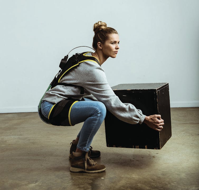 A woman wearing an elaborate harness around her back and thighs squatting while lifting a box.