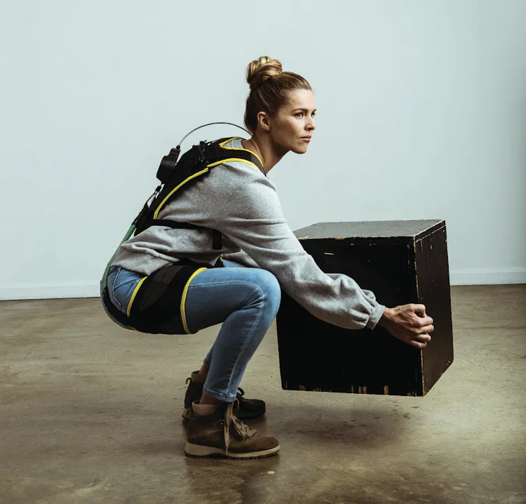 A woman wearing an elaborate harness around her back and thighs squatting while lifting a box.