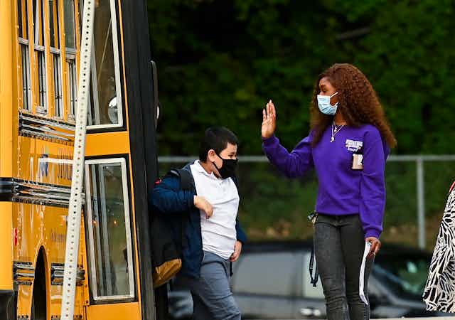 An adult in face mask greets a student getting off a bus.
