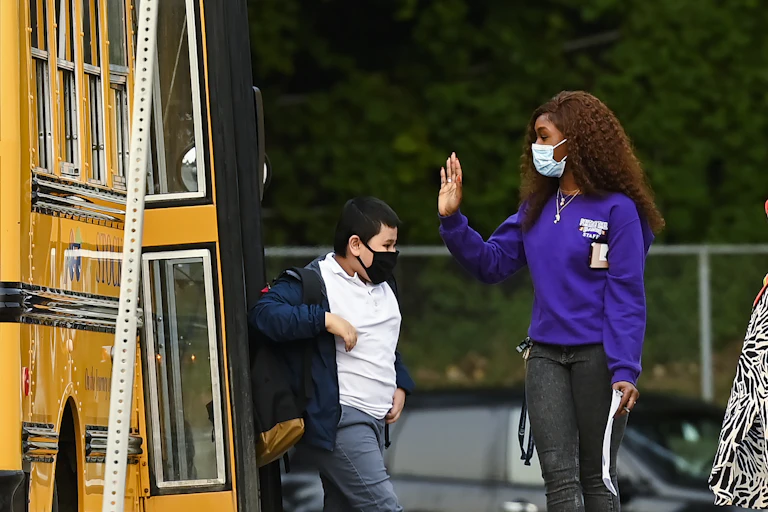 An adult in face mask greets a student getting off a bus.