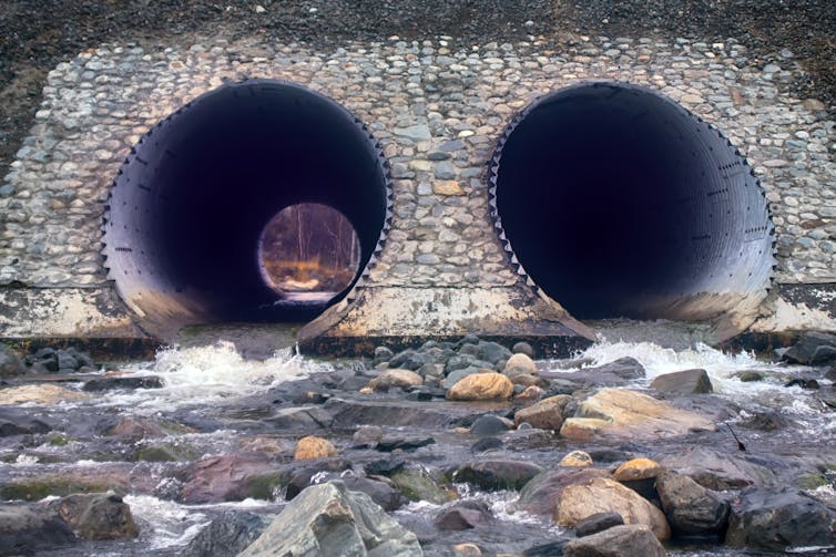 Two large, metal tunnels with flowing water set in the side of a motorway embankment.