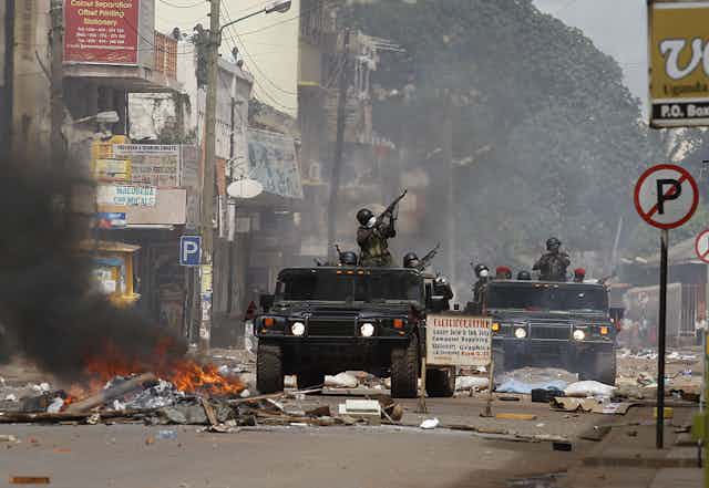 Soldiers point guns while riding military vehicles down a street where a pile of material is burning