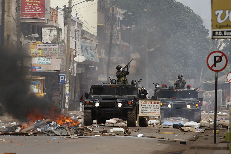 Soldiers point guns while riding military vehicles down a street where a pile of material is burning