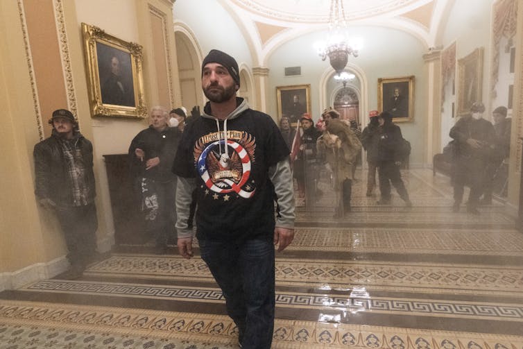 A man wearing a shirt with a large Q on it stands in front of a group in a smoke-filled corridor