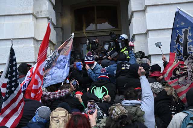 A crowd of people, some holding flags and some holding phones, press several police officers against a door