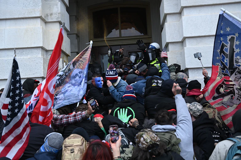A crowd of people, some holding flags and some holding phones, press several police officers against a door