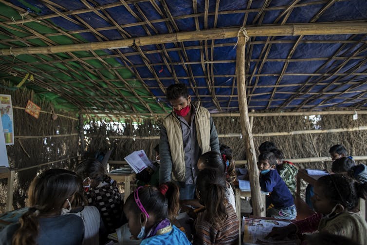 Man in facemask supervises children under a makeshift hut