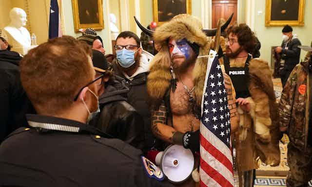 Man wearing fur hat with horns, carrying US flag, surrounded by rioters in the US Capitol Building, January 6 2021.