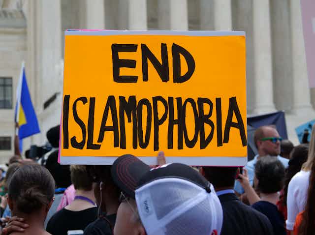 A neon orange sign saying END ISLAMOPHOBIA is held above a crowd