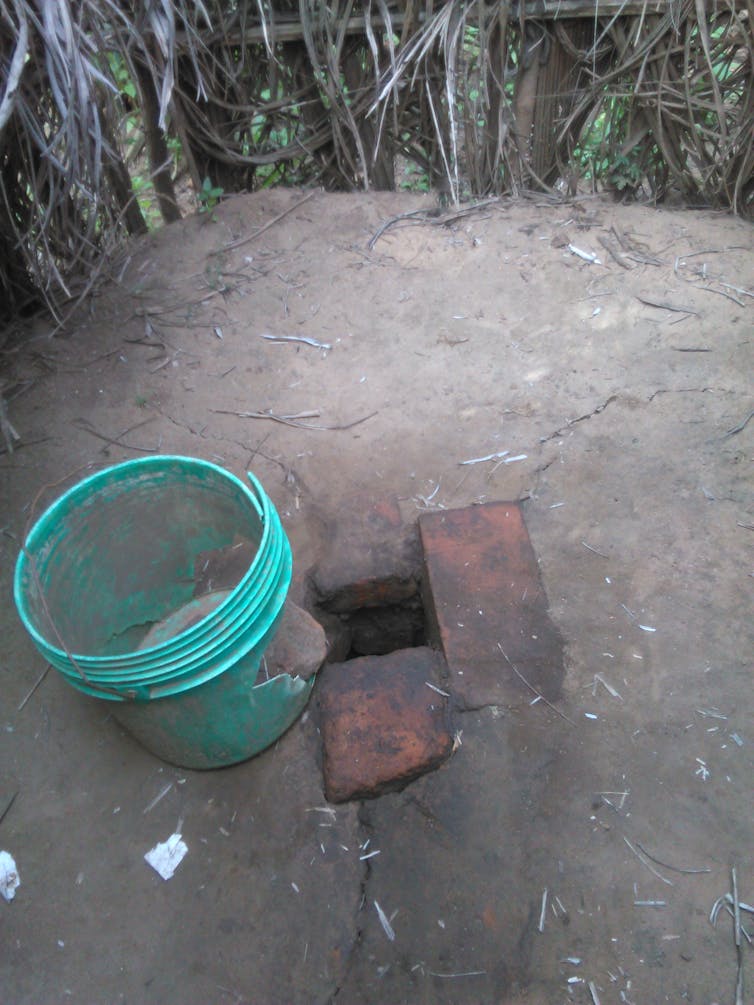 A hole in the ground, lined with two bricks, and a blue bucket beside it