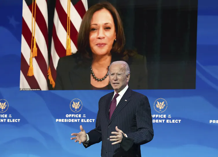 Joe Biden speaks from a stage with Kamala Harris on video screen behind him.