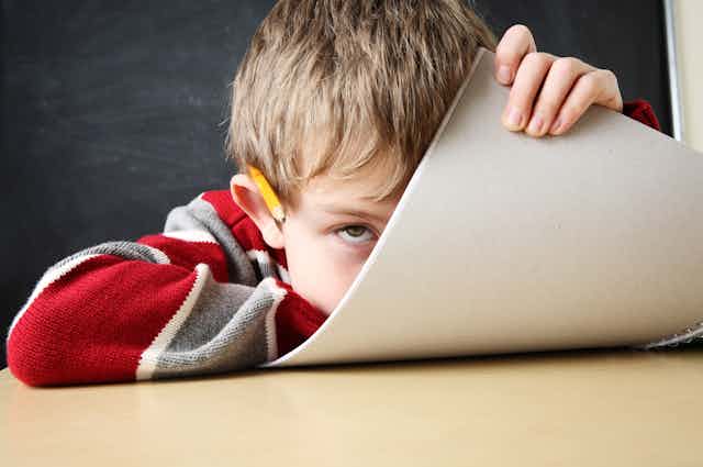 Un niño con cara aburrida, un lapicero sobre la oreja mira a cámara mientras se tapa media cara con un folio.