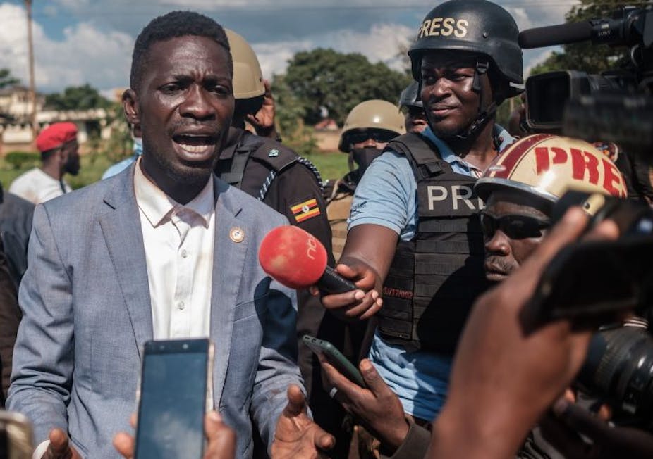 Robert Kyagulanyi speaks to journalists holding a microphone and cellphones and wearing hard hats and protective vests