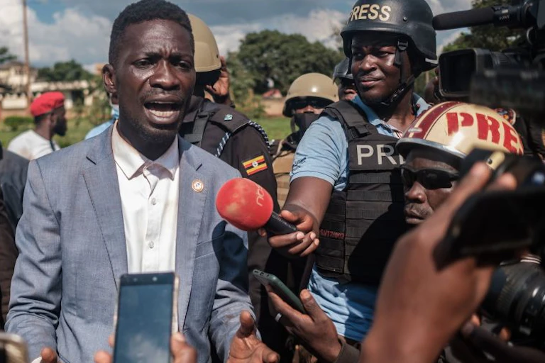 Robert Kyagulanyi speaks to journalists holding a microphone and cellphones and wearing hard hats and protective vests