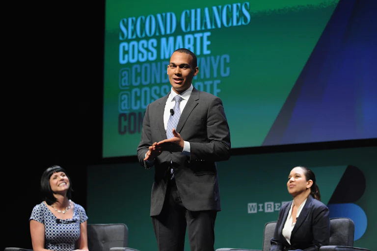A man in a suit and tie speaks while standing between two seated women.