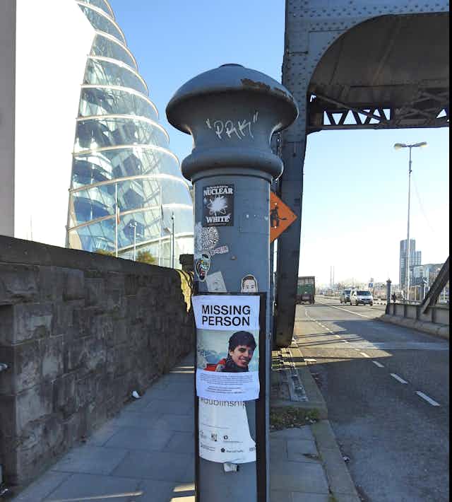 Poster of a missing person on a metal pillar in the middle of a street with a glass building and a metal bridge in the background