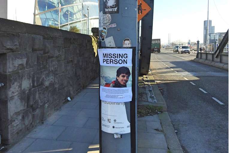 Poster of a missing person on a metal pillar in the middle of a street with a glass building and a metal bridge in the background