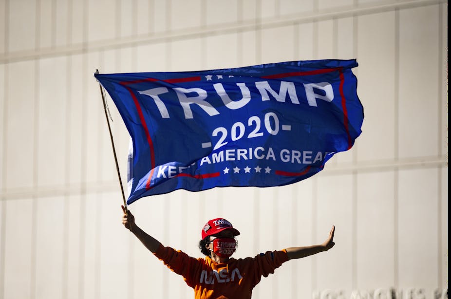Person wearing red Trump hat waves Trump 2020 banner from roof of moving car