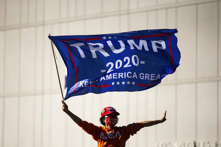 Person wearing red Trump hat waves Trump 2020 banner from roof of moving car