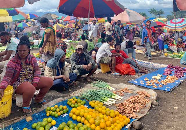A women is sitting next to several men at a market where they are selling a wide range of produce. 