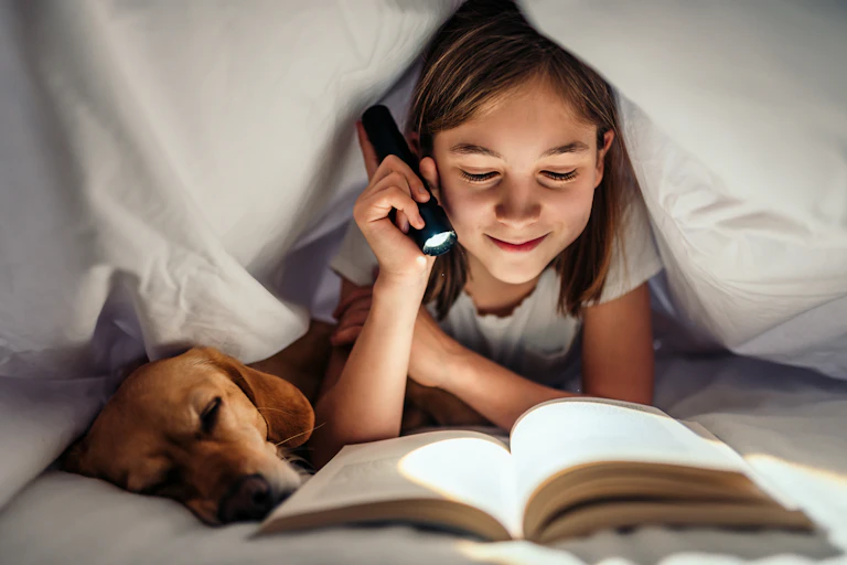A girl reads a book under her bed covers late at night.